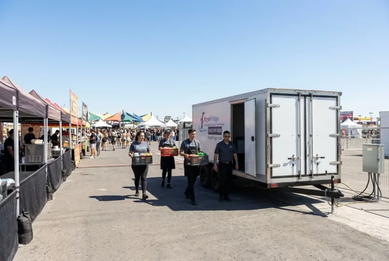 A white refrigerated trailer staged behind a row of festival food booths on a closed-off tree-lined avenue on a sunny afternoon, catering crew in aprons wheeling a hand truck of stacked hotel pans toward the open rear door, crowd and canopy tents blurred in the background
